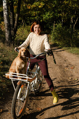 Happy young woman riding a bicycle with her dog in a basket down the road in autumn forest.