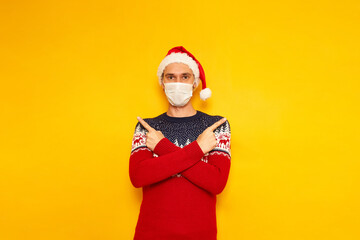 man in Christmas sweater with reindeer, medical mask, red Santa Claus hat points with his index fingers in different directions at an empty place for New Year advertising. isolated yellow background