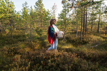 Run away from town: young woman in rubber boots wondering on swamp at sunset searching for berries and mushroom. Joy of relaxing outdoors in beautiful autumn forest. Leisure weekend in nature concept