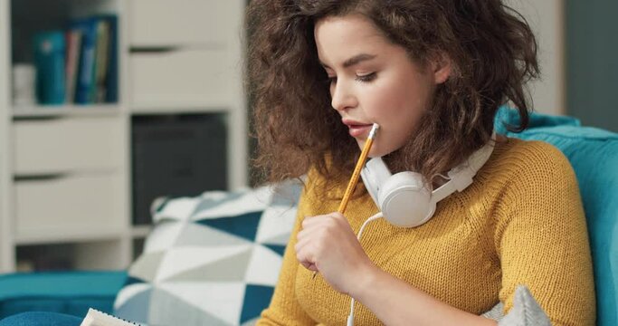 Young beautiful caucasian woman with curly hair in yellow sweater sitting at home on sofa and writing ideas in notebook.