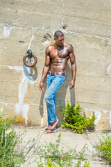 Under the sunshine, a masculine black guy, wearing blue jeans, barefoot and half naked, is standing...