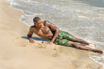 A well built young black guy, half naked, is lying on the beach and playing with waves of ocean..