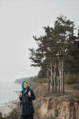 Obraz premium beautiful woman with blue hair in a raincoat and with a backpack stands on a hill above the beach