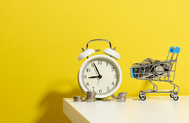 round alarm clock, miniature shopping cart with coins on a white table. Concept time is money,...
