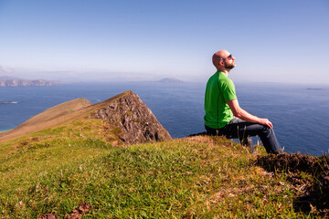 Naklejka na ściany i meble Bald man relaxing and enjoys view on the ocean from a mountain peak, warm sunny day. Male tourist, slim body, wearing green t shirt and blue jeans. Achill island, county Mayo, Ireland.