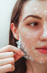 Beautiful young woman with healthy skin removes purifying transparent peel off mask with shiny...