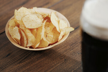 Dark ale in a glass with foam and a wooden small cup with potato chips on a wooden table. Selective focus. Traditional beer and snack