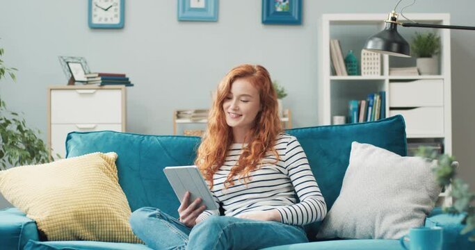 Beautiful cheerful red-haired girl sitting at home on couch and chatting with friends on video call online using tablet.