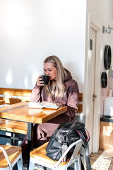 Young woman in a comfortable tracksuit is reading a paper book while drinking coffee in a cafe.