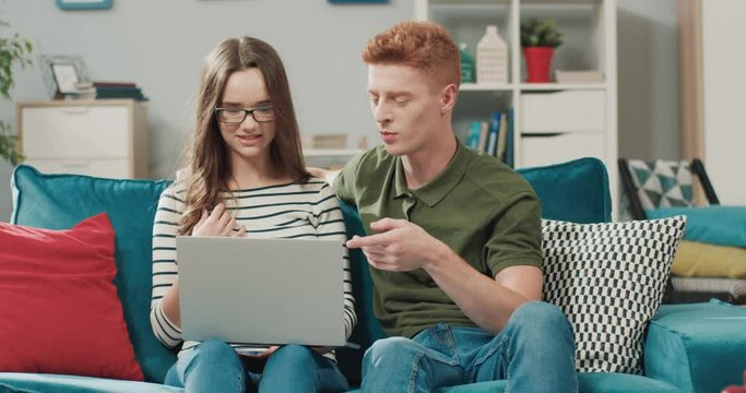 Young beautiful couple sitting at home on couch and spending free time together watching funny videos online on laptop.