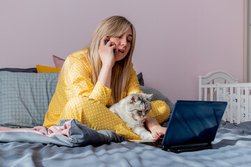 Young woman works on laptop sitting on bed in bedroom next to her pet