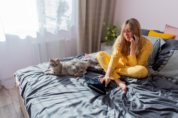 Young woman works on laptop sitting on bed in bedroom next to her pet