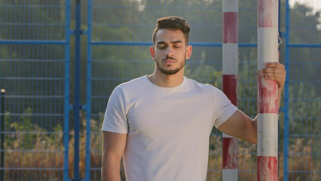 Attractive Hispanic guy in summer sportswear posing at stadium standing at soccer goal. Serious sportsman, coach, goalkeeper or former footballer looking sadly at camera, upset with results of match