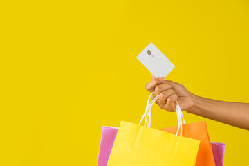 Mano de una mujer con bolsas de compra y una tarjeta para pagar en un fondo amarillo - obrazy, fototapety, plakaty