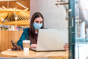 View through glass of young woman in protective mask is working on computer while drinking coffee.