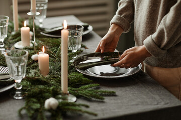 Fototapeta na wymiar Side view close up of unrecognizable woman setting up dining table decorated for Christmas with fir branches and candles in grey tones, copy space