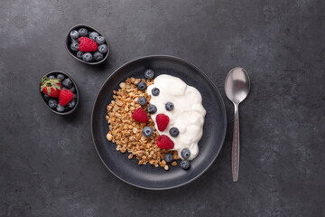 Homemade granola with yogurt and fresh berries in black ceramic plate on dark stone background. Healthy vegan breakfast - 462340773