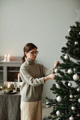 Vertical waist up portrait of young woman decorating Christmas tree at home in elegant silver and...