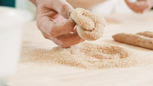 Bakery chef dipping raw dough pieces in white sesame seeds, covering knotted bread rolls in sesame seeds