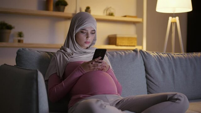 Young pensive pregnant muslim lady sitting on sofa and reading news or baby care instruction online on smartphone