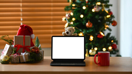 Computer tablet, coffee cup and gift boxes on wooden table near Christmas tree at home.