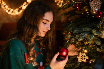 Cute teenager girl decorates christmas tree with red balls in green sweater with deer. Cozy magical...