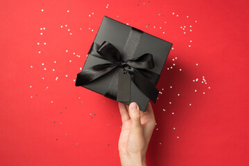 First person top view photo of hand holding black giftbox with black ribbon bow and tag sequins on isolated red background with copyspace