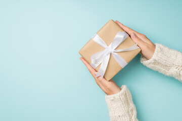 First person top view photo of hands in white knitted pullover holding stylish craft paper giftbox with white ribbon bow on isolated pastel blue background with copyspace
