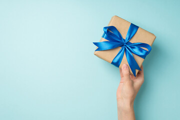 First person top view photo of hand holding craft paper giftbox with vivid blue satin ribbon bow on isolated pastel blue background with copyspace