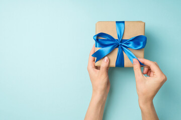 First person top view photo of hands unpacking craft paper giftbox with blue ribbon bow on isolated pastel blue background with copyspace