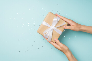 First person top view photo of hands giving craft paper giftbox with white ribbon bow over sequins on isolated pastel blue background with empty space