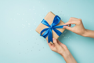 First person top view photo of hands unpacking craft paper gift box with blue satin ribbon bow over shiny sequins on isolated pastel blue background with blank space