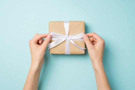 First person top view photo of hands tying white satin ribbon bow on craft paper giftbox on isolated pastel blue background
