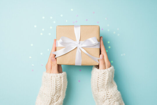 First person top view photo of hands in white sweater holding craft paper giftbox with white ribbon bow over shiny snowflakes confetti on isolated pastel blue background
