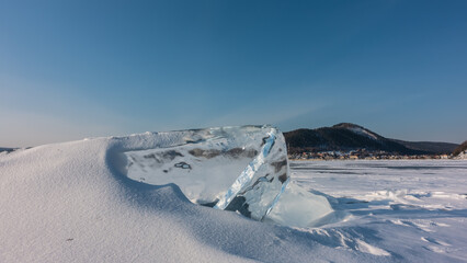 A thick transparent ice floe on a frozen lake. The hummock is partially covered with snow. In the distance, city houses are visible on the shore. A mountain range against a blue sky. Baikal