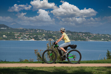 The movement of an electric cyclist along the coastline in the background lake, mountains, blue sky with clouds. Woman on a sports mountain electric bike lake garda.