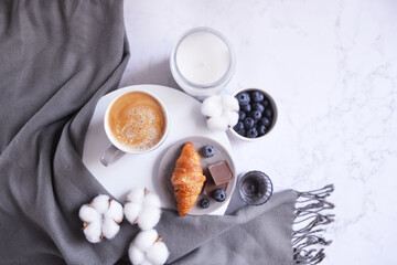 Cup of coffee with croissant, chocolate bar, blueberries, cotton flowers and grey scarf on the white marble background. Autumn, winter coffee break. Cozy warm home latte. Empty place for text. 
