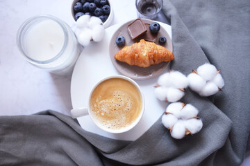 Cup of coffee with croissant, chocolate bar, blueberries, cotton flowers and grey scarf on the white marble background. Autumn, winter coffee break. Cozy warm home latte. Relax at home.