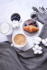 Fototapeta na wymiar Cup of coffee with croissant, chocolate bar, blueberries, cotton flowers and grey scarf on the white marble background. Autumn, winter coffee break. Cozy warm home latte. Relax at home.