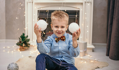 Naklejka premium A Caucasian little boy in a blue shirt and a tiger bow tie is sitting near the fireplace in a room with Christmas balls