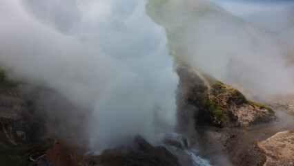 A column of thick steam rises from the cauldron of an erupting geyser, splashes of boiling water scatter around. The hillside is shrouded in fog. Kamchatka. Valley of geysers.