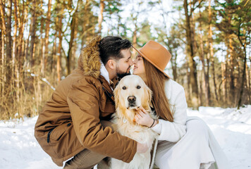 Family outdoors. Romantic lovely woman and handsome man kissing in winter forest having a leisure time with golden retriever dog. - obrazy, fototapety, plakaty