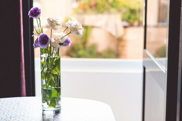 White and purple flowers stand in a vase of water on the table. There is a beautiful view from the window.