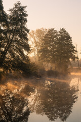 Fototapeta premium Jesienny pejzaż nad wodą. Mgły, drzewa, promienie słońca, rzeka. Staw w Białej na rzece Czerniawce, Gmina Zgierz