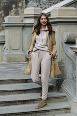 Portrait of fashionable women in beige sports suit, trench coat and stylish suede loafer posing on...