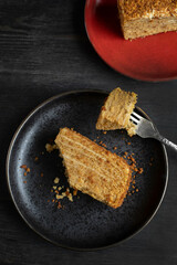 Vegan honey cake on a black plate, metal fork with a piece of cake, another piece of cake on a red plate aside. Black wooden backround. Top view.