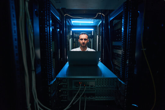 Data Center Worker Sitting At Laptop Before Server Cabinet