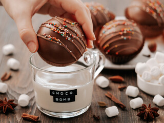 Chocolate cocoa bomb in hand near glass cup with plant-based milk and choco bomb text. Ball made...