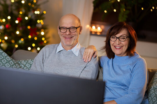 Winter Holidays, Leisure And People Concept - Happy Smiling Senior Couple Watching Tv At Home In Evening Over Christmas Tree Lights On Background