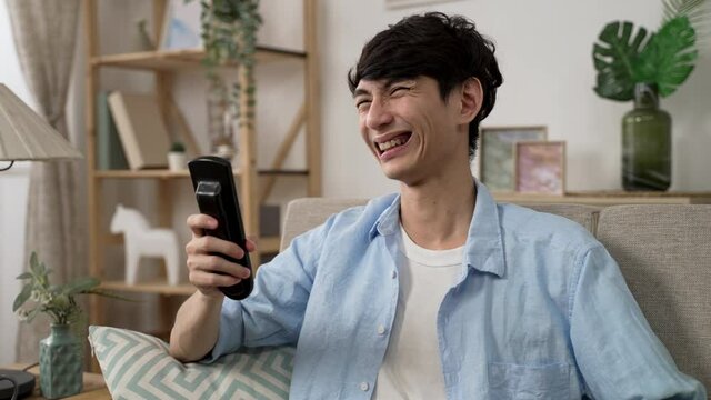 closeup of a cheerful asian male holding remote control is bursting out laughing while watching a comedy on tv in the living room at home